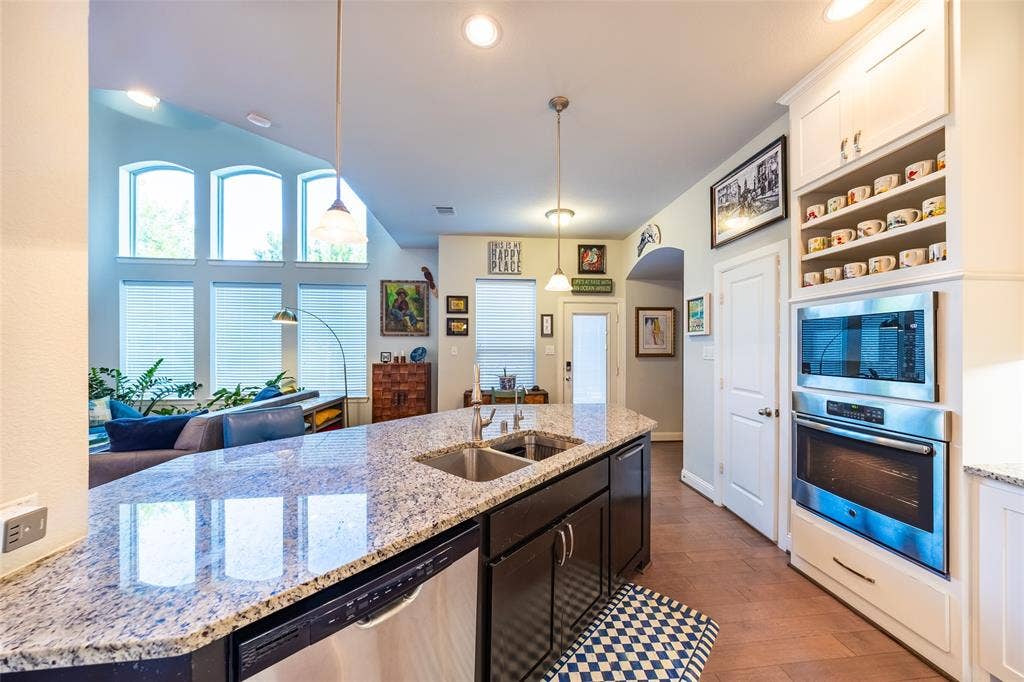 Open-concept living area featuring a kitchen island with dual sinks and granite-finish countertop, stainless steel dishwasher, and built-in wall oven and microwave