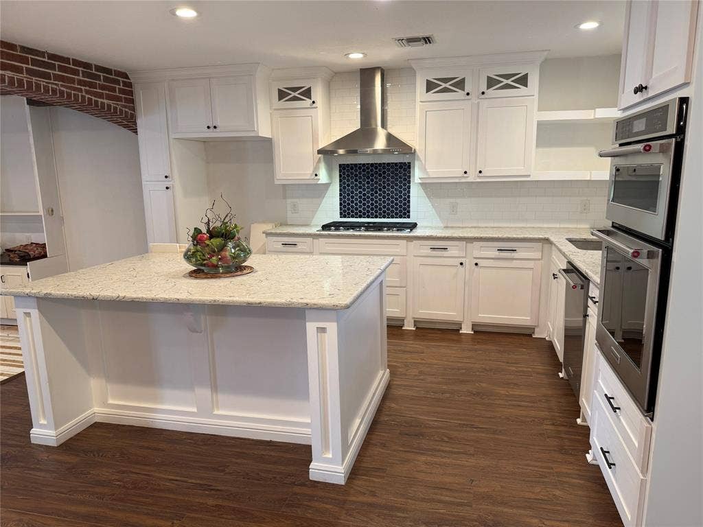 13430 Challaburton Drive, Farmers Branch, TX 75234 Modern kitchen featuring a central island with a light-toned countertop, white cabinetry with dark hardware, a subway tile backsplash, stainless steel appliances including a built-in double oven, and wood-finish flooring