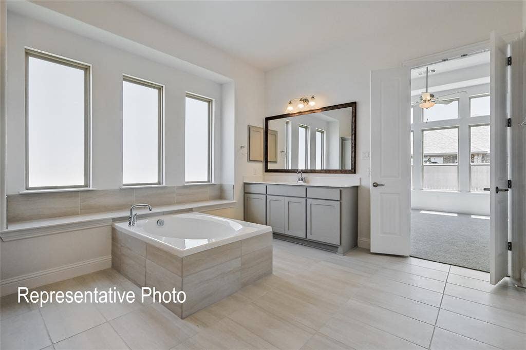 Spacious bathroom featuring a built-in soaking tub with tiled surround, three vertical windows, a dual-sink vanity with light-colored cabinetry, and large format tile flooring