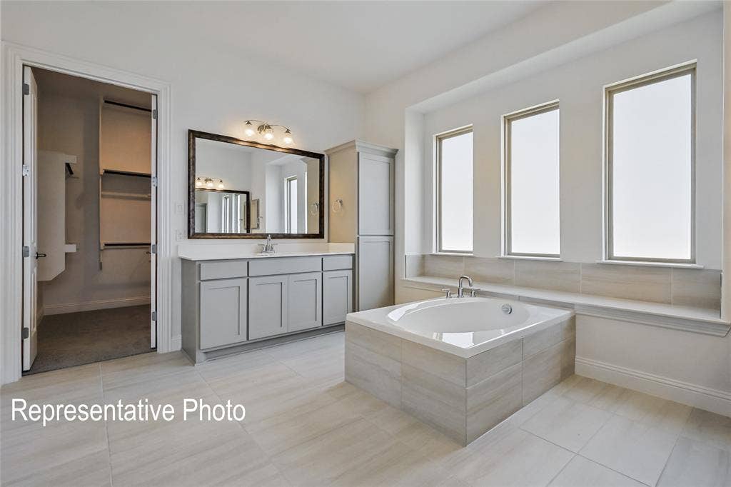 Spacious bathroom featuring a built-in soaking tub with tiled surround, triple rectangular windows, a vanity with ample counter space, and a large framed mirror