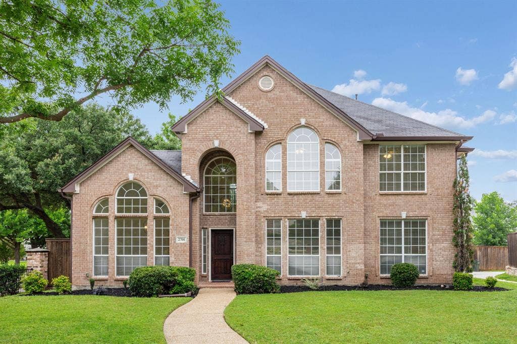 2701 Oates Drive, Plano, TX 75093 Brick exterior with architectural arched windows, a dark wood entry door, a stone walkway, and mature landscaping