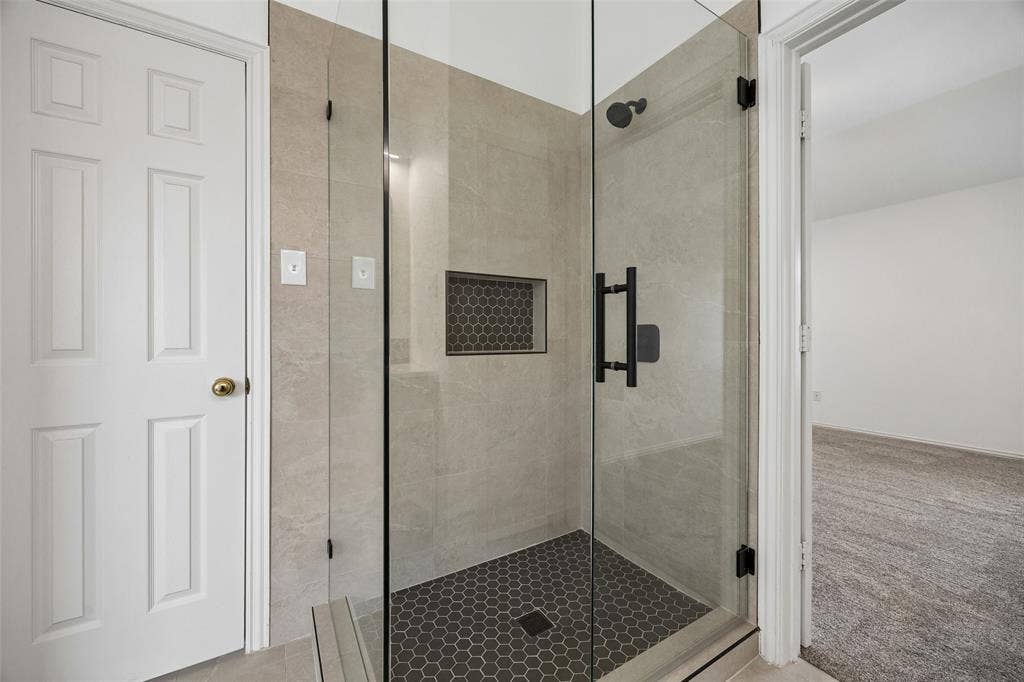 2701 Oates Drive, Plano, TX 75093 Contemporary walk-in shower featuring clear glass enclosure, matte black fixtures, light-toned stone-finish wall tiling, dark hexagonal floor tiling, and a recessed hexagonal tile niche