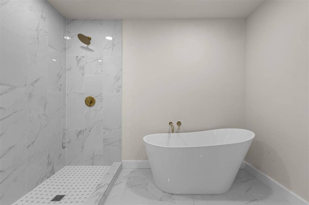 1507 E Waco Avenue, Dallas, TX 75216 Freestanding soaking tub with wall-mounted brass fixtures, marble-look tile flooring, and a walk-in shower featuring matching marble-look wall tiles and brass fixtures