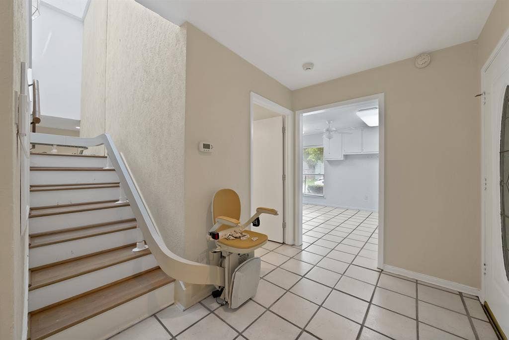 12574 Burninglog Lane, Dallas, TX 75243 Entry foyer featuring white square tile flooring with contrasting grout, a wood-finish staircase with white risers and railing, an integrated stair lift system, and a front entry door with an oval glass insert