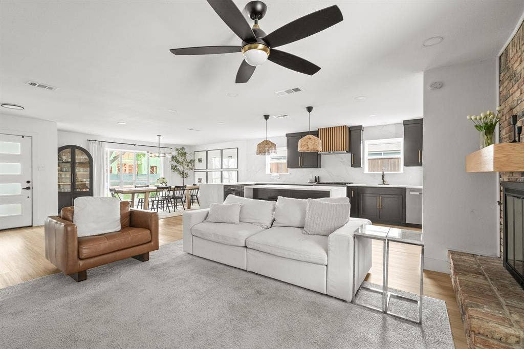 Open-concept living area featuring light wood-finish flooring, a contemporary ceiling fan, a brick fireplace with a wood mantle, a modern kitchen with dark cabinetry, and white countertops