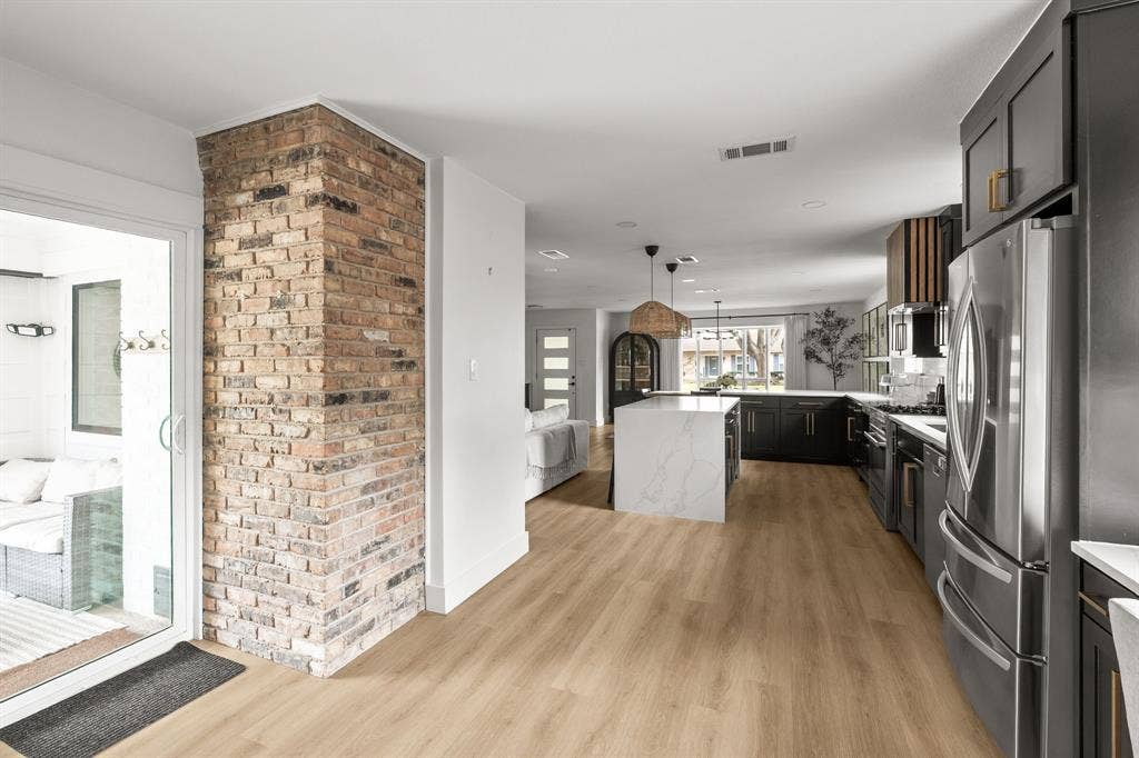 Open-concept kitchen and living area featuring wood-finish flooring, a brick accent wall, a large kitchen island with a waterfall countertop, dark cabinetry, and stainless steel appliances