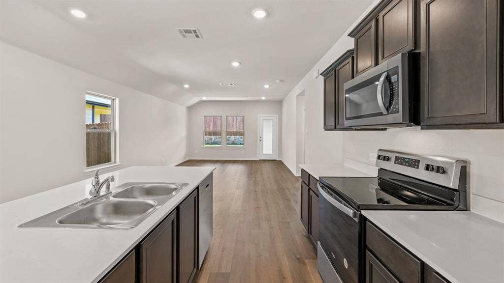 1631 Blaire Drive, Tyler, TX 75702 Open-concept kitchen featuring an island with a double stainless steel sink, dark wood cabinetry, white countertops, and stainless steel appliances, including an electric range and microwave, all complemented by wood-finish flooring and recessed lighting
