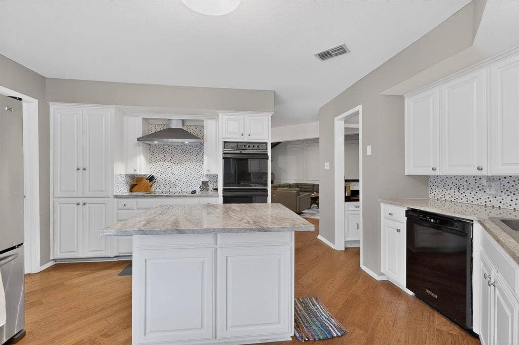 1151 Lynn Way, Waxahachie, TX 75165 Open-concept kitchen featuring an island with a light-toned countertop, white cabinetry, wood-finish flooring, stainless steel range hood, and integrated black appliances