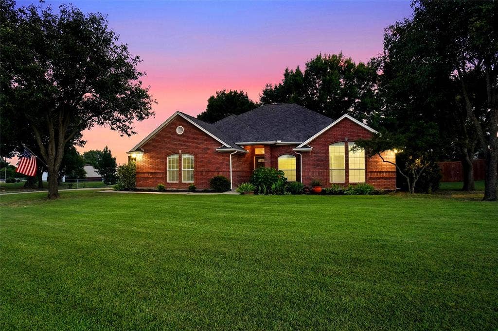 Brick exterior residence featuring a dark shingle roof, mature trees, and a spacious green lawn