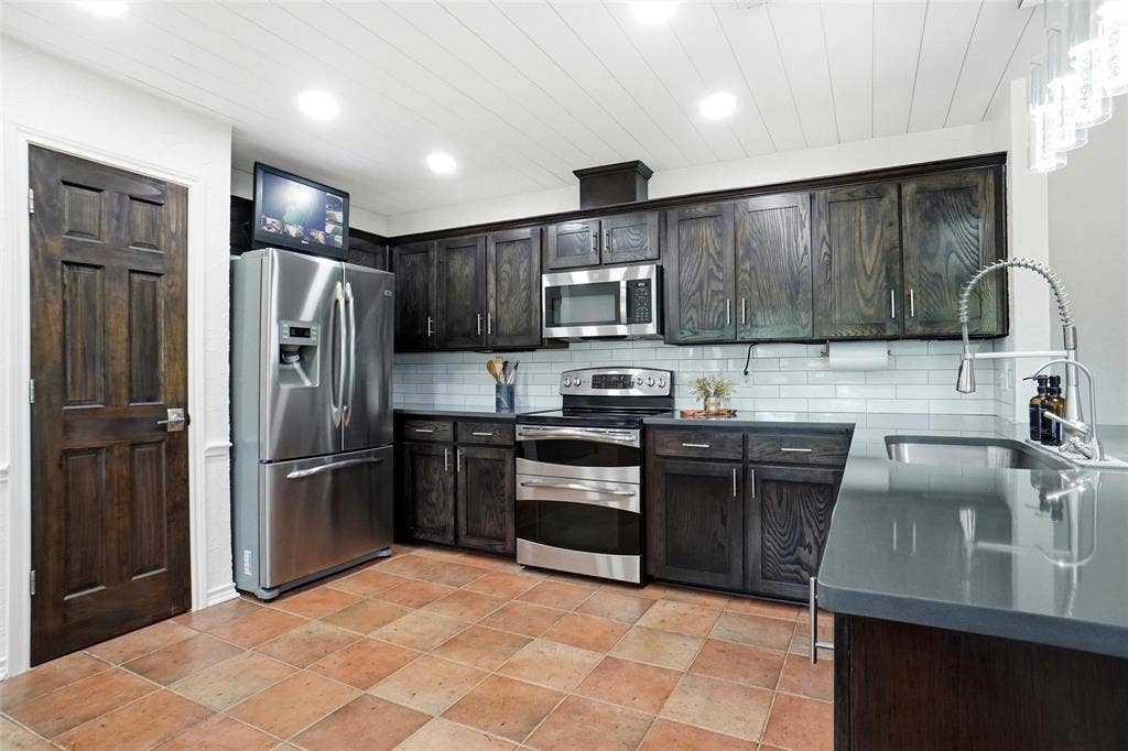 Kitchen featuring dark wood cabinetry, pantry, stainless steel appliances, a subway tile backsplash, quartz countertop, and a large single basin sink with a gooseneck faucet