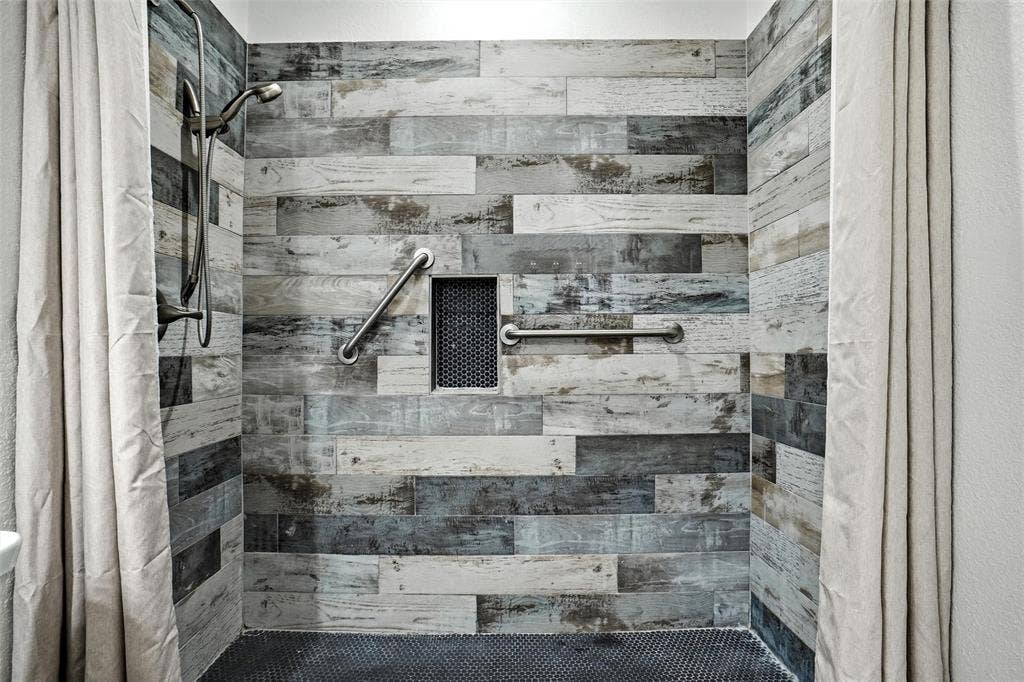 Shower enclosure featuring wood-look tile walls, black penny tile flooring, a recessed shower niche, and two grab bars