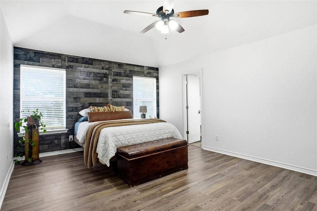 Primary bedroom with a vaulted ceiling, ceiling fan, accent wall with horizontal wood paneling, two windows with blinds, and LVP wood-style flooring