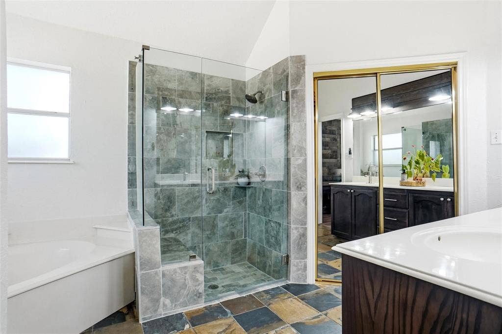 Spacious bathroom featuring a glass-enclosed shower with stone tile surround, corner soaking tub, dual vanity with dark wood cabinetry, and multi-toned slate-style floor tiling