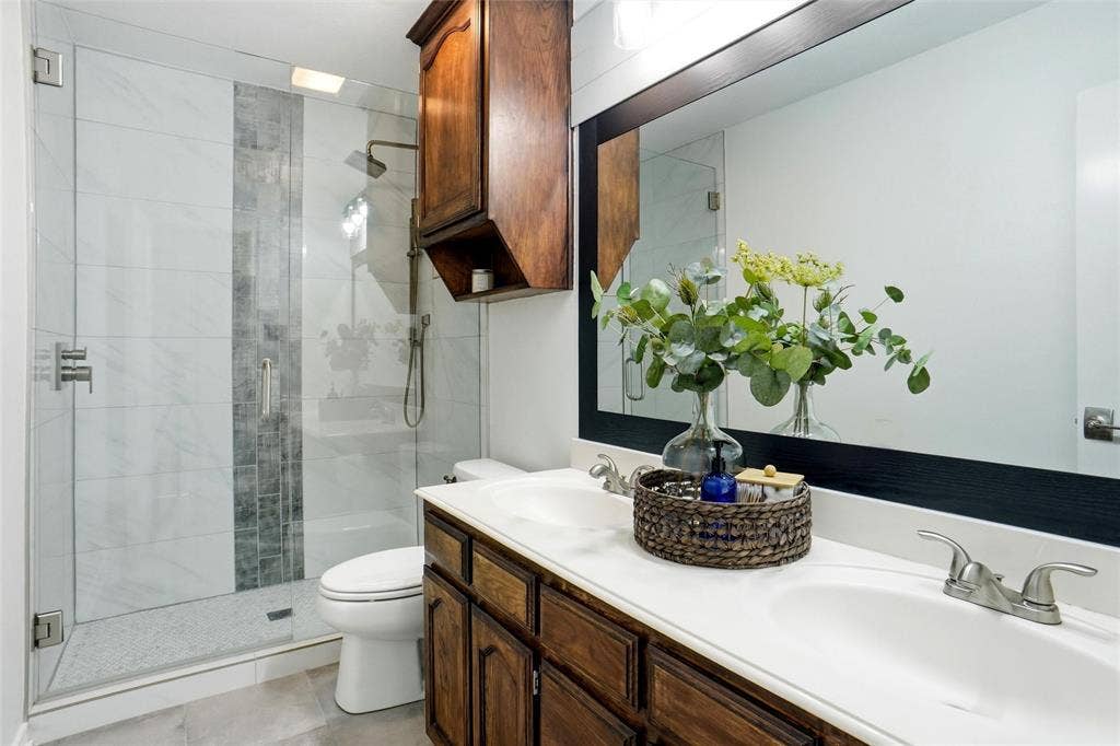 Bathroom featuring a glass-enclosed shower with white and grey tile, vanity with dual sinks, wood-finish cabinetry, and a large wall-mounted mirror
