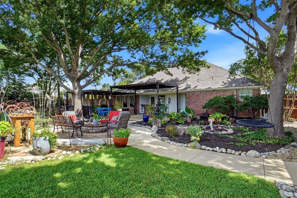Spacious backyard featuring mature trees, a stone-edged fire pit area, a perimeter walkway, brick exterior, and a covered patio area