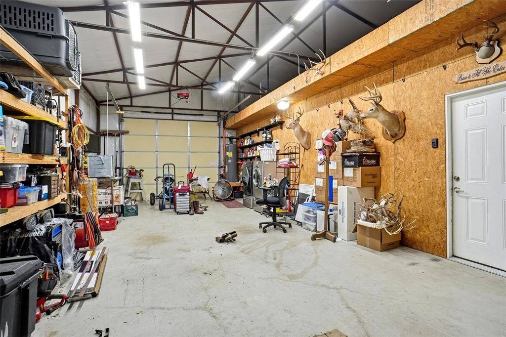 Spacious workshop featuring front and back garage doors, exposed metal beam ceiling, concrete flooring, built-in shelving, and a paneled interior door