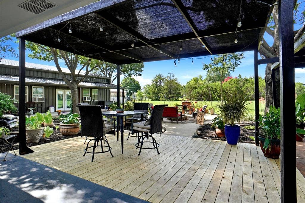 Expansive outdoor living area featuring a wood-finish deck, covered patio with string lighting, concrete patio extension, and mature landscaping