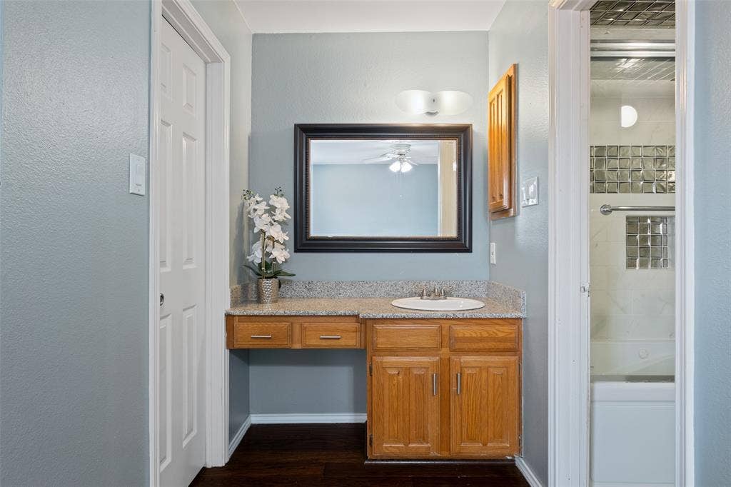 4130 Proton Drive, #47B, Addison, TX 75001 Secondary bathroom vanity area with granite countertop, warm wood cabinetry, a large framed mirror, and a peek into the shower/tub combo beyond.