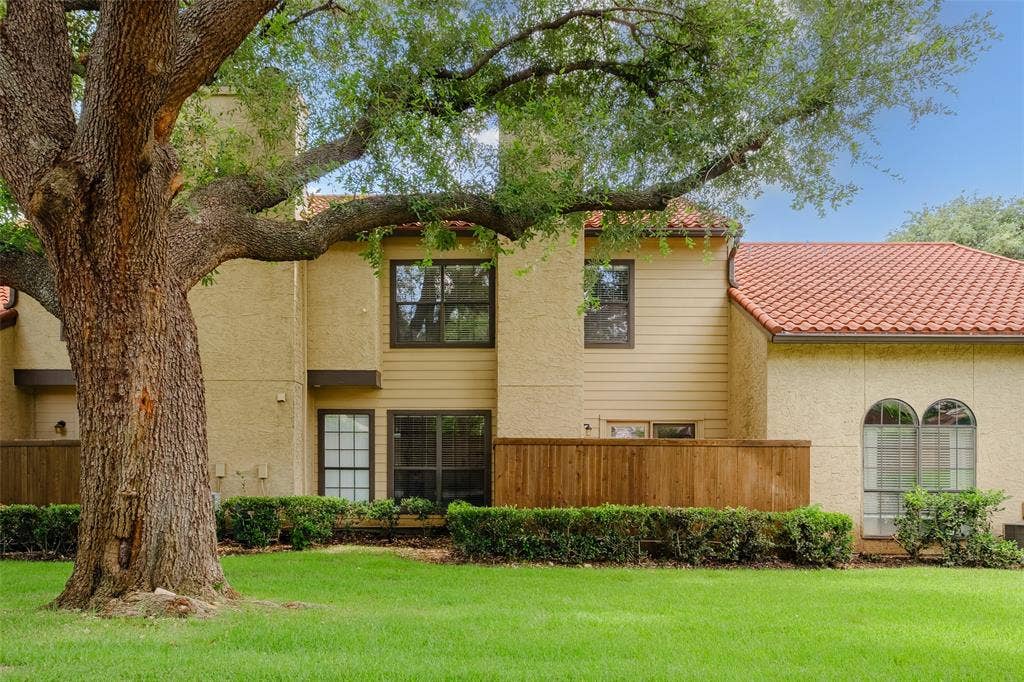 4130 Proton Drive, #47B, Addison, TX 75001 Back exterior view showcasing the clay tile roof, private wood fence patio enclosure, mature live oak canopy, and lush community greenspace.