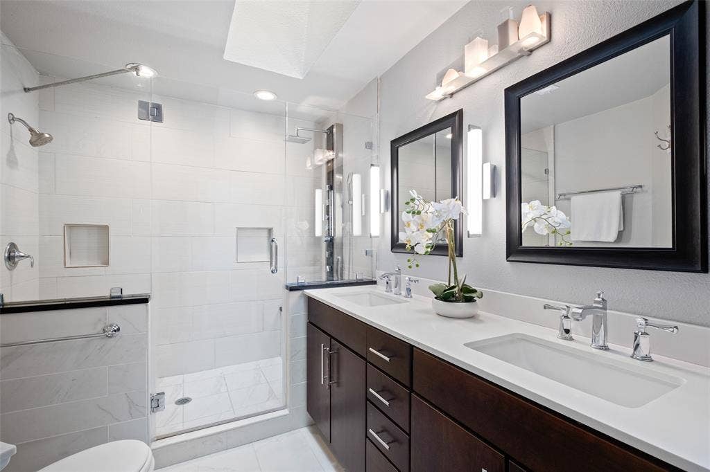 4130 Proton Drive, #47B, Addison, TX 75001 Fully renovated primary bath featuring a double-sink vanity with quartz countertops, dark espresso cabinetry, framed mirrors, modern sconce lighting, and a frameless glass walk-in shower with large-format white tile.