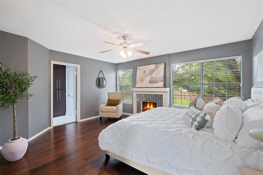 4130 Proton Drive, #47B, Addison, TX 75001 Spacious primary bedroom with hardwood floors, a second wood-burning fireplace with tile surround, multiple windows overlooking the greenery, ceiling fan, and direct access to the primary bath and walk-in closet.