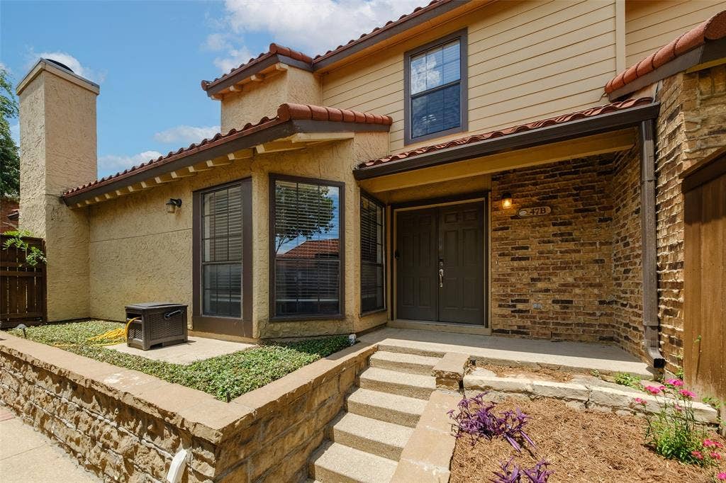 4130 Proton Drive, #47B, Addison, TX 75001 Close-up of the covered front entry with brick surround, tile roof overhang, unit signage, and blooming landscape plantings framing the steps — a welcoming and private arrival experience.