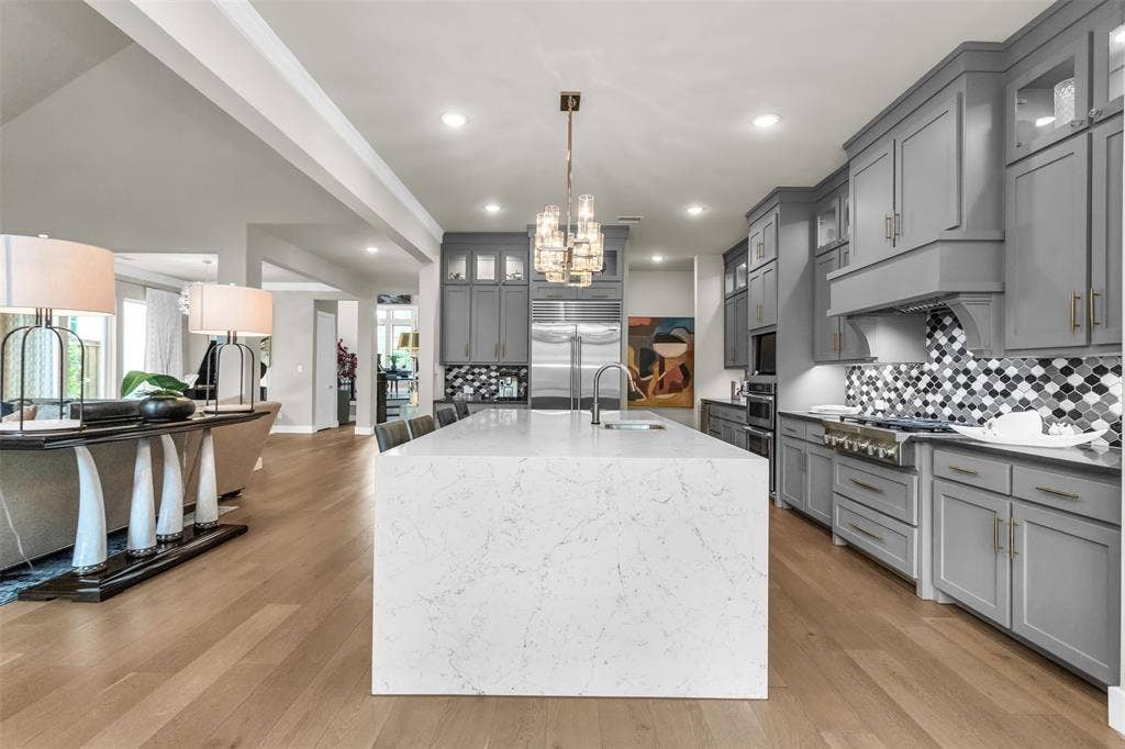 8138 Edgefield Court, Frisco, TX 75035 Gourmet kitchen featuring a waterfall island with a built-in sink, gray cabinetry, a decorative range hood, stainless steel appliances, and wood-finish flooring