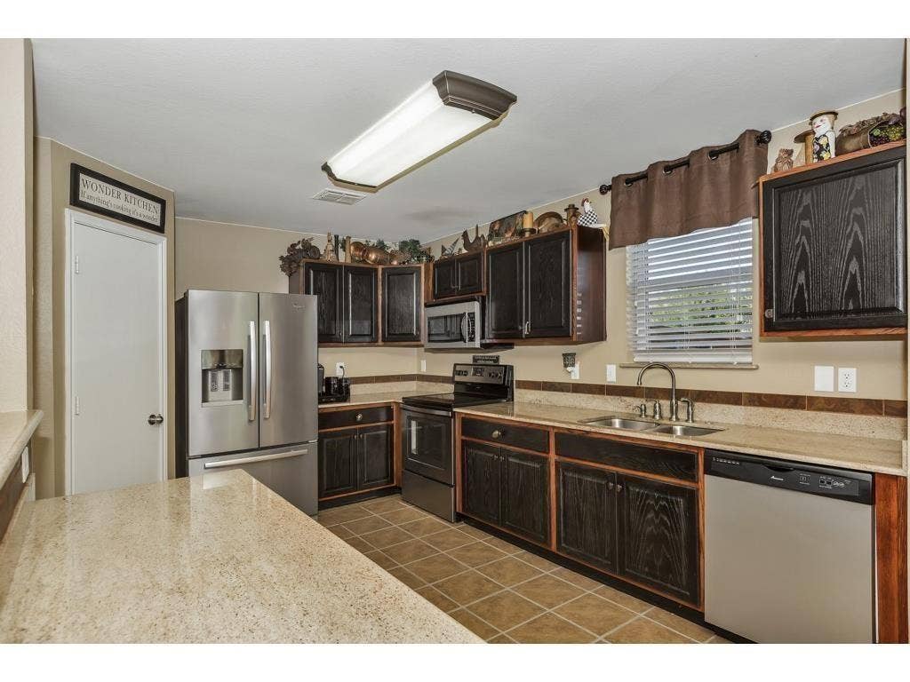2912 Lake Terrace Drive, Wylie, TX 75098 Kitchen featuring dark wood-finish cabinetry, light speckled countertops, a double basin stainless steel sink, and stainless steel appliances including a refrigerator and dishwasher