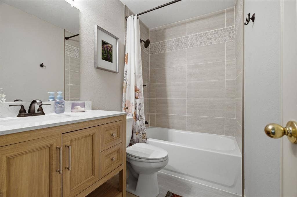 628 Cardinal Lane, Keller, TX 76248 Bathroom featuring a wood-finish vanity with a white countertop, a bronze-finish faucet, a toilet, and a shower-over-tub with light-toned rectangular tile and a mosaic accent band