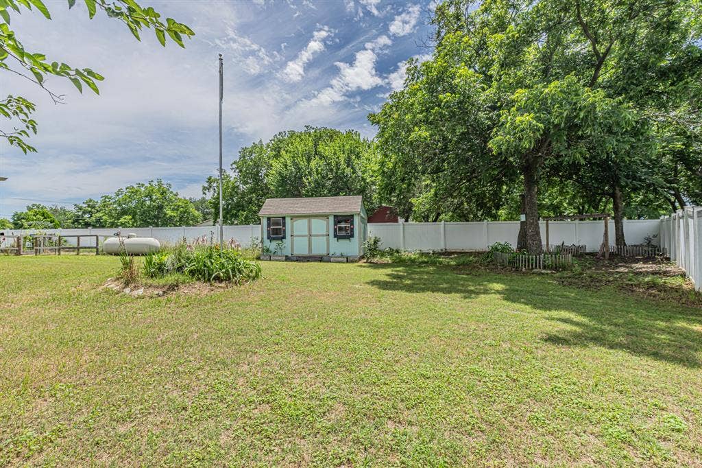 204 S Spring Branch Trail, Springtown, TX 76082 Expansive backyard featuring a manicured lawn, established trees, a light blue storage shed with black shutters, a white privacy fence, and a central flagpole