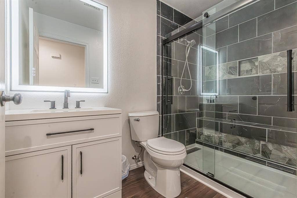 204 S Spring Branch Trail, Springtown, TX 76082 Bathroom featuring a white vanity with integrated sink, rectangular LED-lit mirror, dark wood-finish flooring, a commode, and a shower with dark tiled walls, a built-in bench, and clear glass enclosure