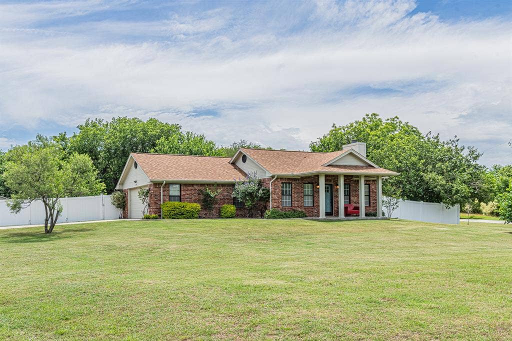 204 S Spring Branch Trail, Springtown, TX 76082 Brick exterior residence featuring a covered front porch with white columns, a two-car garage, and a white privacy fence encompassing a portion of the property