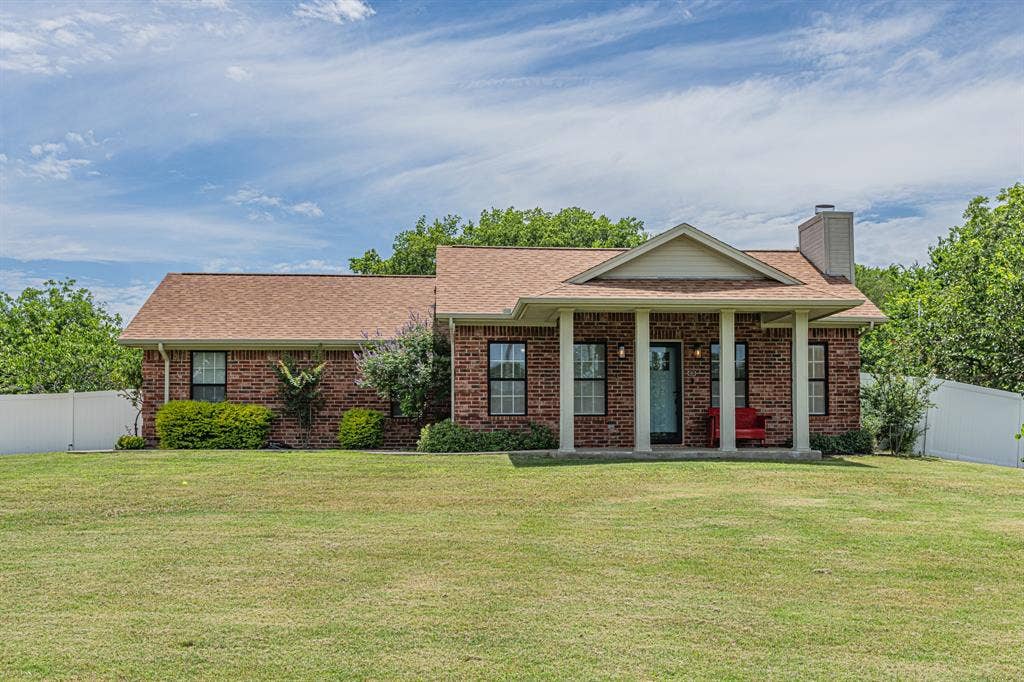 204 S Spring Branch Trail, Springtown, TX 76082 Brick facade residence featuring a covered front porch with column supports, a tan shingle roof, a prominent chimney, and mature landscaping