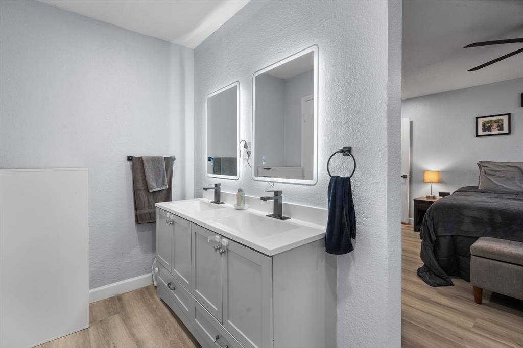 7629 Cedarhill Road, Watauga, TX 76148 Dual vanity with integrated sinks and white countertops, illuminated mirrors, matte black fixtures, and wood-finish flooring