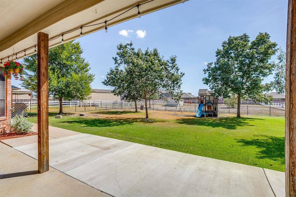 Covered patio with string lighting, overlooking a green lawn with mature trees and a fenced perimeter