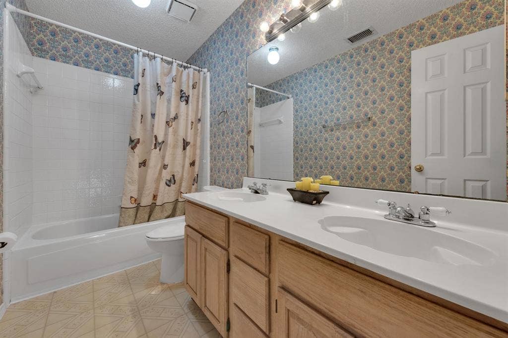 4622 Devonshire Drive, Grand Prairie, TX 75052 Bathroom featuring a dual vanity with light wood cabinetry and white countertops, a large mirror with strip lighting, a white bathtub with tile surround, and patterned wallpaper