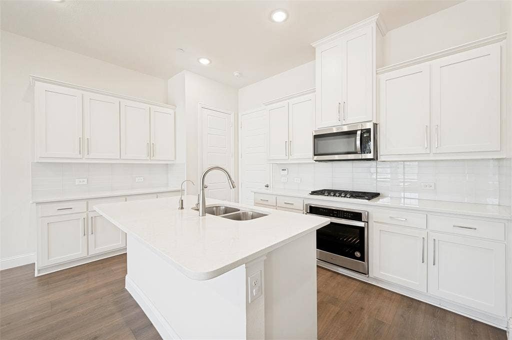 12986 Jk Ojha Street, Farmers Branch, TX 75234 Modern kitchen featuring a central island with an undermount double-basin sink, white cabinetry, stainless steel appliances, white subway tile backsplash, and wood-finish flooring