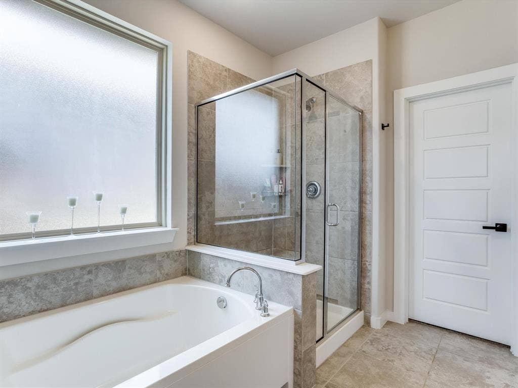 1021 Daylily Drive, Northlake, TX 76226 Bathroom featuring a built-in soaking tub with chrome fixtures, a clear glass enclosed shower with a built-in niche, light-toned tile flooring, and a modern white paneled door with matte black hardware