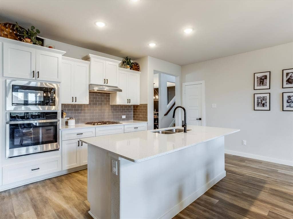 1021 Daylily Drive, Northlake, TX 76226 Kitchen featuring white cabinetry, stainless steel appliances including a built-in microwave and oven, a gray subway tile backsplash, a center island with a sink and matte black faucet, and wood-finish flooring
