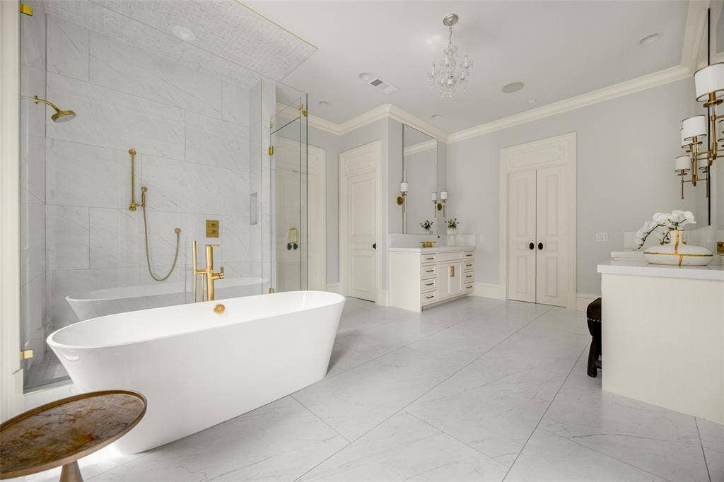 Spacious bathroom featuring a freestanding soaking tub, an expansive glass-enclosed shower with gold-finish fixtures, dual vanities with integrated sinks, and large-format tile flooring