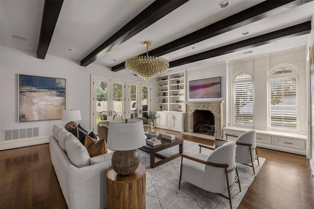 Living space featuring dark wood ceiling beams, a carved stone fireplace, extensive built-in shelving, hardwood flooring, and French doors leading to an outdoor area