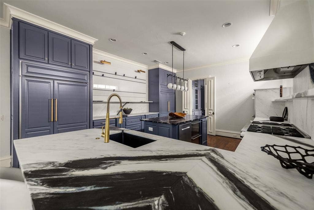 Kitchen featuring a large island with a black sink and gold-finish faucet, stone countertops with prominent veining, integrated paneled refrigerator, built-in range with a large hood, and blue cabinetry with gold-finish hardware