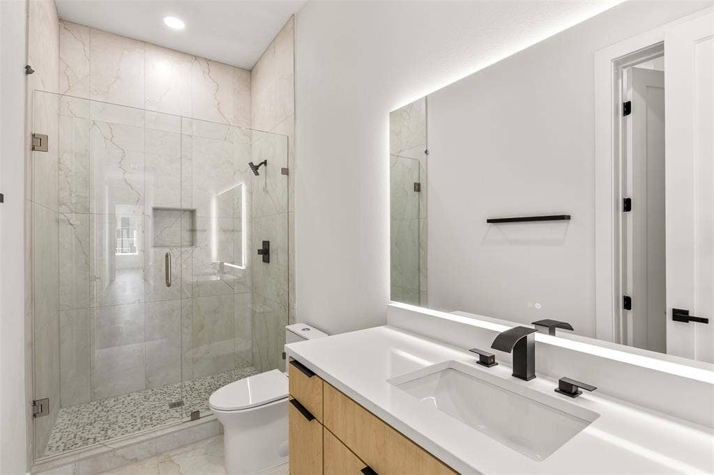 1407 Pueblo Street, Dallas, TX 75212 Modern bathroom featuring a frameless glass shower enclosure, large format tile walls, pebble-finish shower floor, an illuminated mirror, and a wood-finish vanity with a white countertop and matte black fixtures