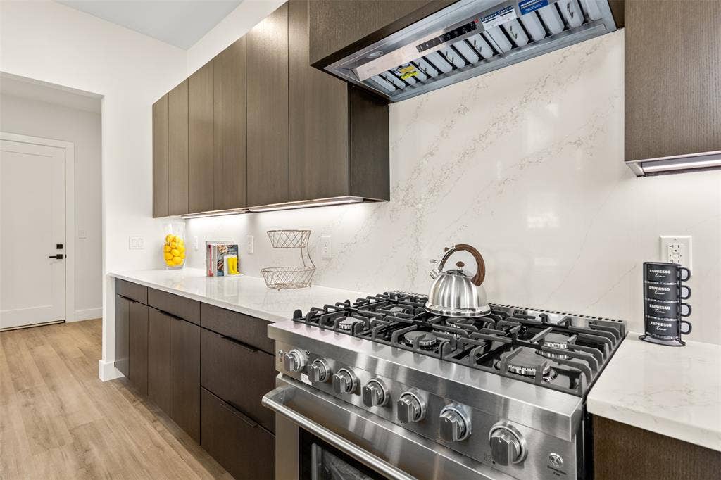 1407 Pueblo Street, Dallas, TX 75212 Gourmet kitchen featuring a stainless steel gas range, coordinating range hood, dark wood-finish cabinetry, light stone countertops, and a full-height stone slab backsplash