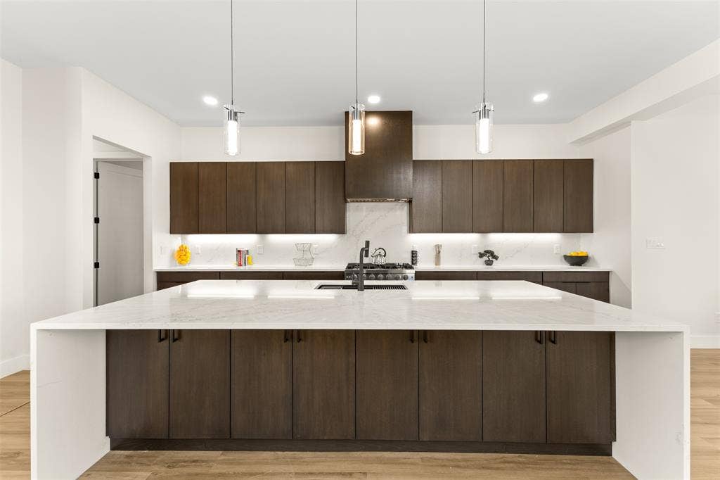1407 Pueblo Street, Dallas, TX 75212 Kitchen featuring a large waterfall island with a light-toned countertop, dark wood-finish cabinetry, a gas range with an overhead vent hood, a light-toned backsplash, and recessed and pendant lighting