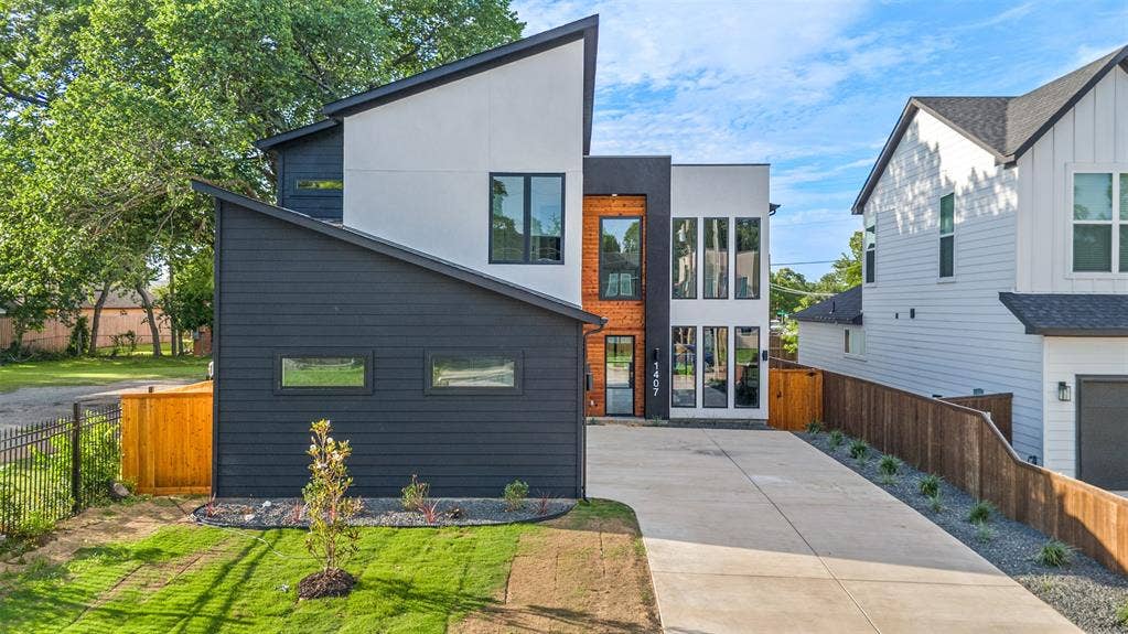 1407 Pueblo Street, Dallas, TX 75212 Modern architectural facade featuring contrasting dark and white siding, an asymmetrical roofline, a prominent multi-pane window array, a wood-accented entryway, and a concrete driveway
