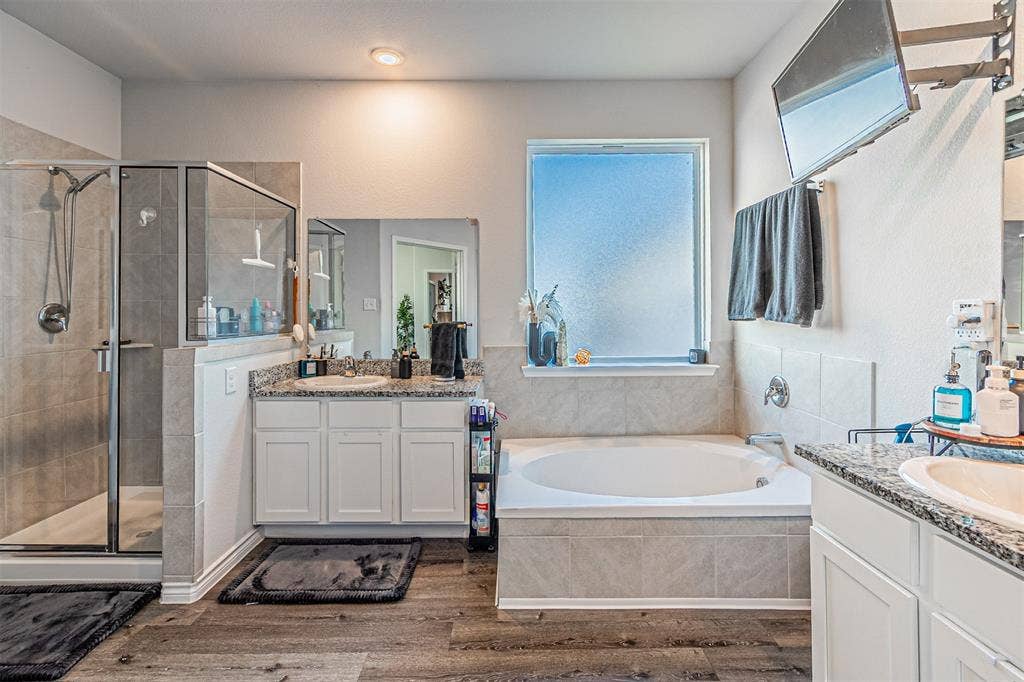 Spacious bath featuring wood-finish flooring, a glass-enclosed shower with tile surround, a built-in soaking tub with tile surround, and dual vanities with stone countertops