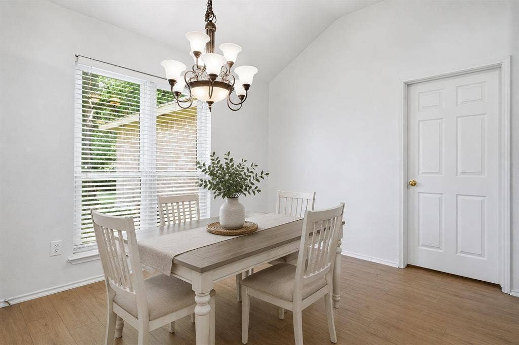 Vaulted ceiling dining area featuring wood-finish flooring, a contemporary chandelier, and a large window with horizontal blinds. *Photo is Virtually staged using AI.
