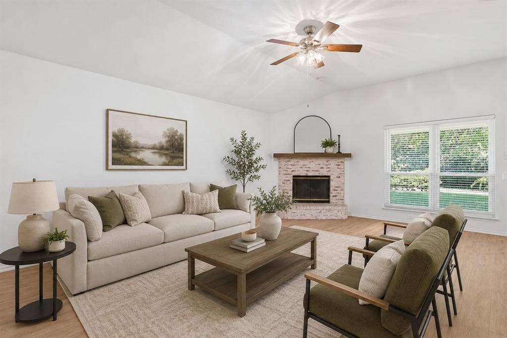 Vaulted ceiling with a ceiling fan, a brick fireplace with a wood mantel, wood-finish flooring, and large windows.*Photo is virtually staged using AI.