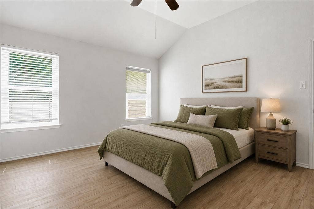 Vaulted ceiling bedroom with wood-finish flooring, two large windows with blinds, and a ceiling fan. *Photo virtually staged using AI
